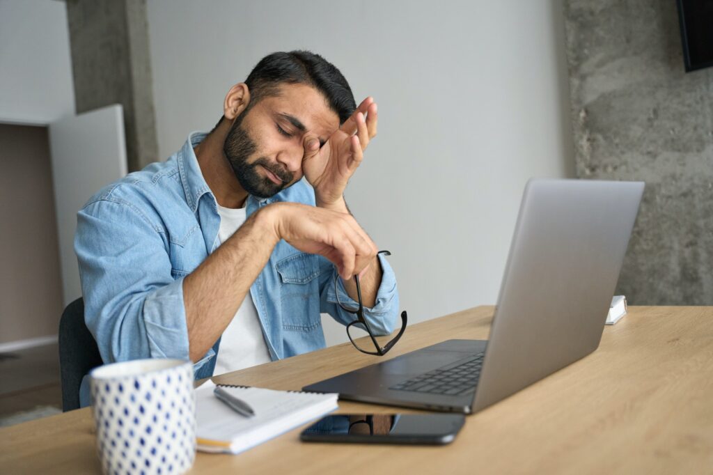 Young tired Hispanic indian student rubbing eyes sitting at desk with laptop.